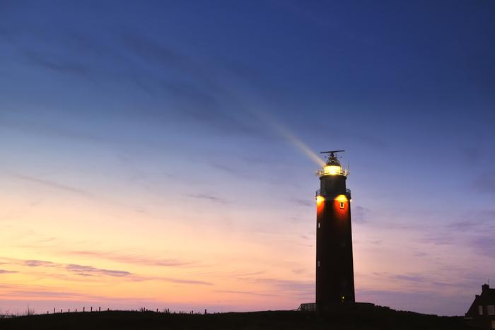 A lighthouse beams light across a twilight sky, silhouetted against soft pink and deep blue hues.