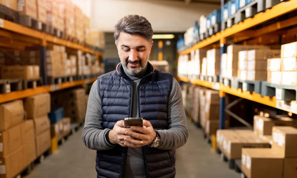 A man stands in a warehouse surrounded by boxes, holding a cell phone, representing secure access to unmanaged devices.