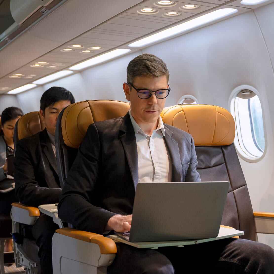A man in a suit sits on an airplane, working on a laptop, illustrating the complexities of VDI in remote work environments.